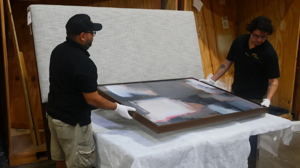 Two men carefully handling a large abstract painting on a table.